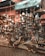 A vibrant market stall filled with an array of brass and copper lamps and ornaments, intricately designed and hanging in various shapes and sizes. The background shows a rustic wall, and the foreground has a cobblestone pathway. The atmosphere is rich and bustling with crafted metalwork that glints under soft lighting.