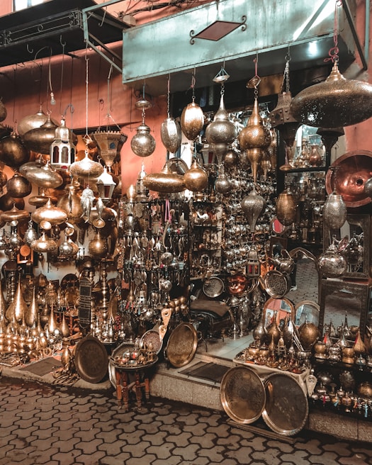 A vibrant market stall filled with an array of brass and copper lamps and ornaments, intricately designed and hanging in various shapes and sizes. The background shows a rustic wall, and the foreground has a cobblestone pathway. The atmosphere is rich and bustling with crafted metalwork that glints under soft lighting.