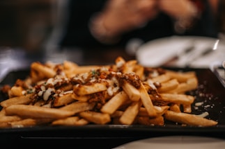 A plate of crispy golden French fries topped with melted cheese and a savory sauce, garnished with herbs. The focus is on the fries, while the background is softly blurred, indicating a dining setting.