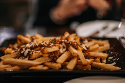 A plate of crispy golden French fries topped with melted cheese and a savory sauce, garnished with herbs. The focus is on the fries, while the background is softly blurred, indicating a dining setting.