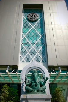A large building facade with a modern geometric design features a prominent statue of a figure with multiple arms, located in front of the entrance. The figure sits in a meditative pose, framed by an arched structure. The overall architectural design includes a mix of glass and metal elements.