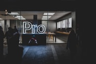 A brightly lit store interior is visible through large glass windows, showcasing a sleek and modern design. The word 'Pro' is prominently displayed in glowing white neon lights near the entrance. Inside, there are tables with electronic devices on display, and the area appears clean and organized. Outside, silhouettes of people can be seen, some standing and some seated on the ground.