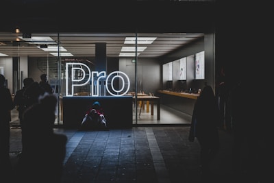 A brightly lit store interior is visible through large glass windows, showcasing a sleek and modern design. The word 'Pro' is prominently displayed in glowing white neon lights near the entrance. Inside, there are tables with electronic devices on display, and the area appears clean and organized. Outside, silhouettes of people can be seen, some standing and some seated on the ground.