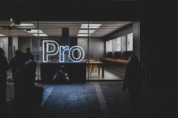 A brightly lit store interior is visible through large glass windows, showcasing a sleek and modern design. The word 'Pro' is prominently displayed in glowing white neon lights near the entrance. Inside, there are tables with electronic devices on display, and the area appears clean and organized. Outside, silhouettes of people can be seen, some standing and some seated on the ground.