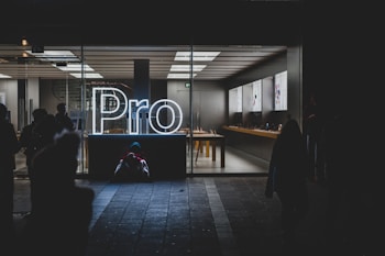 A brightly lit store interior is visible through large glass windows, showcasing a sleek and modern design. The word 'Pro' is prominently displayed in glowing white neon lights near the entrance. Inside, there are tables with electronic devices on display, and the area appears clean and organized. Outside, silhouettes of people can be seen, some standing and some seated on the ground.