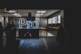 A brightly lit store interior is visible through large glass windows, showcasing a sleek and modern design. The word 'Pro' is prominently displayed in glowing white neon lights near the entrance. Inside, there are tables with electronic devices on display, and the area appears clean and organized. Outside, silhouettes of people can be seen, some standing and some seated on the ground.