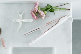 On a light marble surface, there is a white and rose gold flat iron next to a white hair clip. Nearby, pink flowers and green stems are casually arranged, adding a touch of nature. To the side, a few rose gold bobby pins are scattered.
