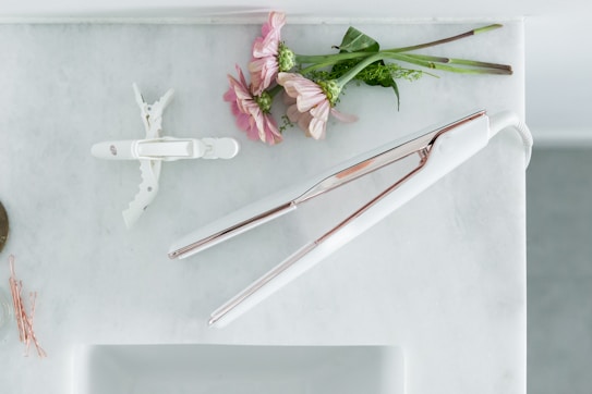On a light marble surface, there is a white and rose gold flat iron next to a white hair clip. Nearby, pink flowers and green stems are casually arranged, adding a touch of nature. To the side, a few rose gold bobby pins are scattered.
