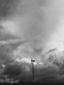 A large wind turbine stands against a dramatic, cloudy sky. The clouds are dense and dark, creating a moody atmosphere. A small airplane is visible high in the sky, adding a sense of scale and contrast to the scene.