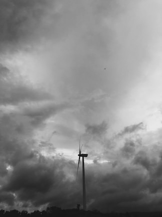 A large wind turbine stands against a dramatic, cloudy sky. The clouds are dense and dark, creating a moody atmosphere. A small airplane is visible high in the sky, adding a sense of scale and contrast to the scene.