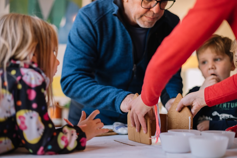 two person making gingerbread house and three children watching them