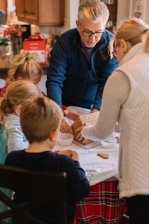 A group of people, including children and an adult, are gathered around a table engaged in building a gingerbread house. The table is covered with a red and white checkered tablecloth, and there are bowls with green candy or icing placed nearby. The adult is guiding the children in assembling the gingerbread pieces.