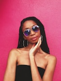 A person with braided hair wearing reflective sunglasses and large hoop earrings stands against a vibrant pink background. The individual has one hand gently touching their face and is dressed in a black, off-the-shoulder top.