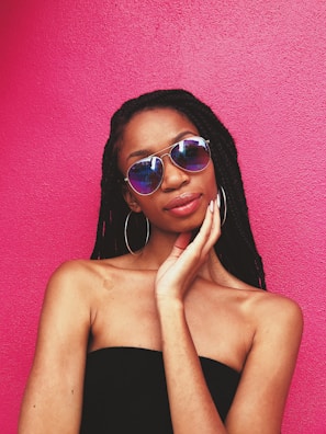 A person with braided hair wearing reflective sunglasses and large hoop earrings stands against a vibrant pink background. The individual has one hand gently touching their face and is dressed in a black, off-the-shoulder top.