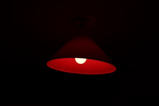 A red lampshade with a glowing light bulb inside, casting a warm, dim light in the surrounding darkness. The lampshade has a conical shape, and the light bulb is centrally positioned, creating a soft halo effect.