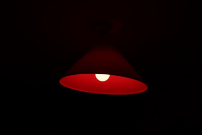 A red lampshade with a glowing light bulb inside, casting a warm, dim light in the surrounding darkness. The lampshade has a conical shape, and the light bulb is centrally positioned, creating a soft halo effect.