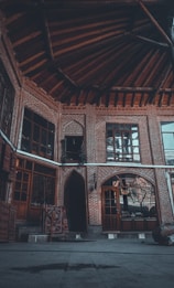 The image features the interior courtyard of a traditional brick building with arched doorways and wooden-framed windows. The architecture displays intricate brickwork patterns and a prominent wooden ceiling with beam structures. Carpets are seen hanging, adding a touch of color and texture to the scene.