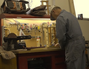 A craftsman wearing a cap and denim coveralls is working at a cluttered workbench. The wall above the bench is lined with various tools such as pliers, screwdrivers, and scissors. A large radio or speaker is placed on the top shelf. The craftsman is engaged in a task that involves precision and focus.