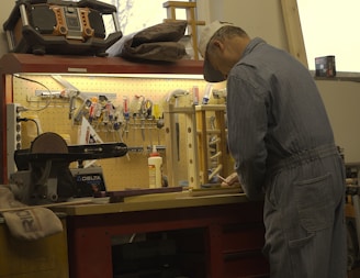 A craftsman wearing a cap and denim coveralls is working at a cluttered workbench. The wall above the bench is lined with various tools such as pliers, screwdrivers, and scissors. A large radio or speaker is placed on the top shelf. The craftsman is engaged in a task that involves precision and focus.