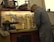 A craftsman wearing a cap and denim coveralls is working at a cluttered workbench. The wall above the bench is lined with various tools such as pliers, screwdrivers, and scissors. A large radio or speaker is placed on the top shelf. The craftsman is engaged in a task that involves precision and focus.
