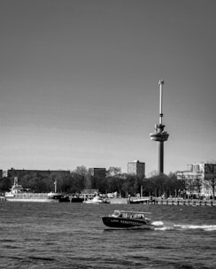 A black and white photograph of a cityscape featuring a prominent tower with an observation deck. A river is in the foreground with a boat that has the name 'SS Rotterdam' on its side. In the background, there are trees and buildings along the riverbank.