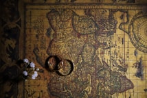 An antique map with intricate details, featuring aged and slightly faded colors. Two interlocking rings are placed on the map, casting a shadow. Small white flowers are arranged to the side, adding a delicate touch.