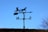 A weathervane silhouette against a clear blue sky, featuring a horse and buggy motif with directional indicators for north and south.
