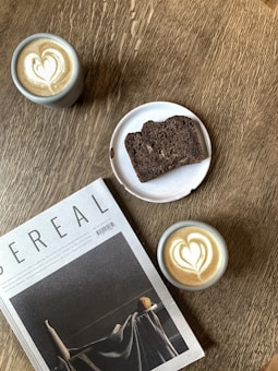 A wooden table features two cups of coffee with heart-shaped latte art, a sliced piece of dark bread on a white plate, and a magazine titled 'Cereal.' The setting creates an inviting and cozy atmosphere.