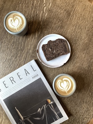 A wooden table features two cups of coffee with heart-shaped latte art, a sliced piece of dark bread on a white plate, and a magazine titled 'Cereal.' The setting creates an inviting and cozy atmosphere.