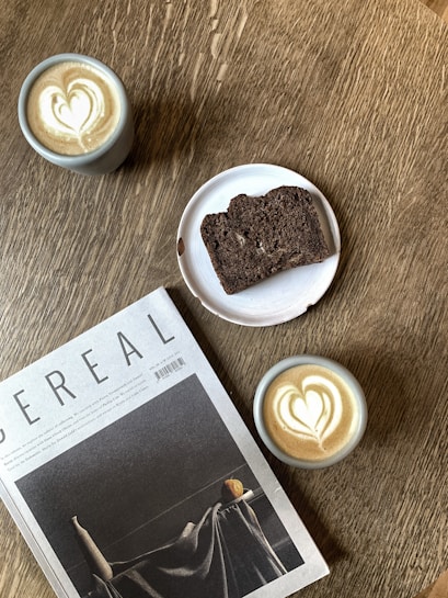 A wooden table features two cups of coffee with heart-shaped latte art, a sliced piece of dark bread on a white plate, and a magazine titled 'Cereal.' The setting creates an inviting and cozy atmosphere.
