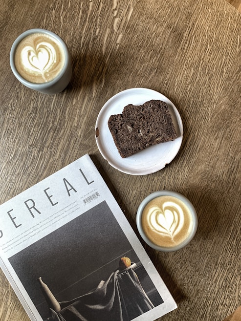 A wooden table features two cups of coffee with heart-shaped latte art, a sliced piece of dark bread on a white plate, and a magazine titled 'Cereal.' The setting creates an inviting and cozy atmosphere.