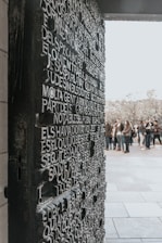 A textured wall or door featuring raised, three-dimensional text in a block-letter style occupies the foreground. The background shows a group of people gathered in an outdoor setting, possibly near a public monument or attraction.