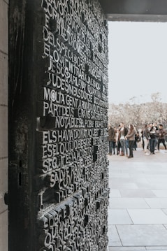 A textured wall or door featuring raised, three-dimensional text in a block-letter style occupies the foreground. The background shows a group of people gathered in an outdoor setting, possibly near a public monument or attraction.