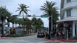A bustling street corner lined with palm trees and a mix of modern and Art Deco architecture. A group of people is waiting at the sidewalk near a restaurant entrance. A tour bus is stopped at a traffic light, and various vehicles are on the road. The atmosphere is lively, typical of a popular tourist area.