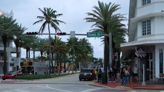 A bustling street corner lined with palm trees and a mix of modern and Art Deco architecture. A group of people is waiting at the sidewalk near a restaurant entrance. A tour bus is stopped at a traffic light, and various vehicles are on the road. The atmosphere is lively, typical of a popular tourist area.