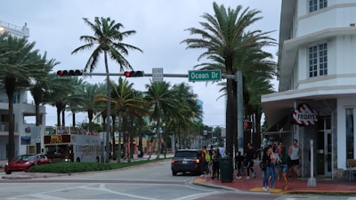 A bustling street corner lined with palm trees and a mix of modern and Art Deco architecture. A group of people is waiting at the sidewalk near a restaurant entrance. A tour bus is stopped at a traffic light, and various vehicles are on the road. The atmosphere is lively, typical of a popular tourist area.