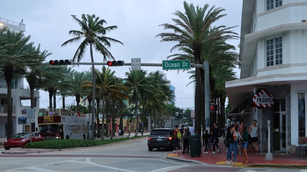 A bustling street corner lined with palm trees and a mix of modern and Art Deco architecture. A group of people is waiting at the sidewalk near a restaurant entrance. A tour bus is stopped at a traffic light, and various vehicles are on the road. The atmosphere is lively, typical of a popular tourist area.