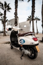 A white scooter with a 'Via Vespa' logo is parked on a sandy area near palm trees, suggesting a coastal or beachside environment. The scooter is facing away from the viewer, with a brown seat and a helmet placed on one of the mirrors. The sky is overcast, adding a calm and relaxed atmosphere to the setting.
