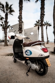 A white scooter with a 'Via Vespa' logo is parked on a sandy area near palm trees, suggesting a coastal or beachside environment. The scooter is facing away from the viewer, with a brown seat and a helmet placed on one of the mirrors. The sky is overcast, adding a calm and relaxed atmosphere to the setting.