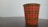 A paper cup with a red and black plaid pattern and yellow accents is placed on a dark wooden surface. The background is a plain, light-colored wall.
