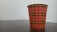 A paper cup with a red and black plaid pattern and yellow accents is placed on a dark wooden surface. The background is a plain, light-colored wall.