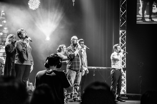 A group of people is on stage, each holding a microphone, presumably performing a song. The setting features stage lights and illuminated decorations. There is a camera operator in the foreground focusing on the performers. The scene captures a concert or a live performance atmosphere.