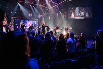 A live music performance takes place on a stage with a band and singers under bright, colorful stage lights. Audience members are standing in a dimly lit venue, watching and enjoying the performance. Decorations are visible, suggesting a festive or concert atmosphere, and a large screen displays visuals and lyrics.