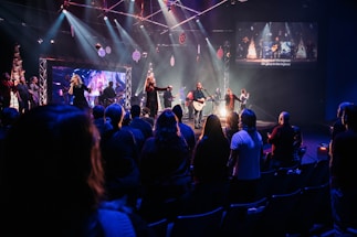A live music performance takes place on a stage with a band and singers under bright, colorful stage lights. Audience members are standing in a dimly lit venue, watching and enjoying the performance. Decorations are visible, suggesting a festive or concert atmosphere, and a large screen displays visuals and lyrics.