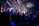 A live music performance takes place on a stage with a band and singers under bright, colorful stage lights. Audience members are standing in a dimly lit venue, watching and enjoying the performance. Decorations are visible, suggesting a festive or concert atmosphere, and a large screen displays visuals and lyrics.