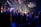 A live music performance takes place on a stage with a band and singers under bright, colorful stage lights. Audience members are standing in a dimly lit venue, watching and enjoying the performance. Decorations are visible, suggesting a festive or concert atmosphere, and a large screen displays visuals and lyrics.