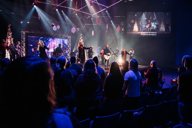 A live music performance takes place on a stage with a band and singers under bright, colorful stage lights. Audience members are standing in a dimly lit venue, watching and enjoying the performance. Decorations are visible, suggesting a festive or concert atmosphere, and a large screen displays visuals and lyrics.