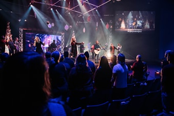 A live music performance takes place on a stage with a band and singers under bright, colorful stage lights. Audience members are standing in a dimly lit venue, watching and enjoying the performance. Decorations are visible, suggesting a festive or concert atmosphere, and a large screen displays visuals and lyrics.