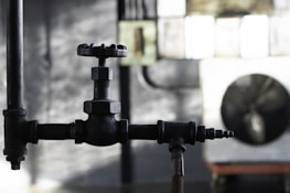 A close-up view of a black metal valve in an industrial setting, with blurred background elements including pipes and a large industrial fan. The light is low, creating a moody and slightly dramatic effect.