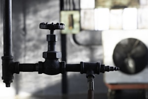 A close-up view of a black metal valve in an industrial setting, with blurred background elements including pipes and a large industrial fan. The light is low, creating a moody and slightly dramatic effect.